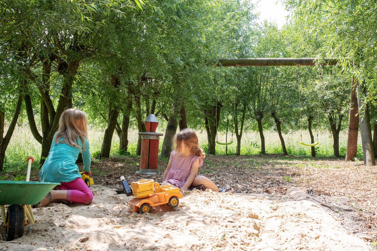 Zwei Kinder spielen im Sandkasten mit Spielzeug in einem grünen, bewaldeten Spielplatz.