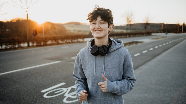 Person joggt auf Straße bei Sonnenuntergang, trägt grauen Hoodie und Kopfhörer um den Hals.