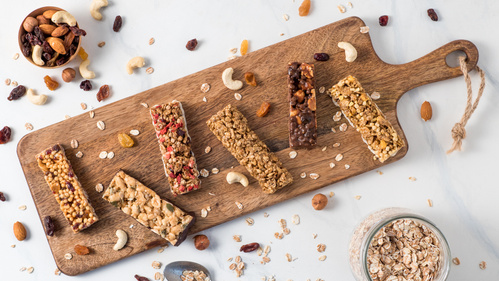 Mehrere Müsli- und Nussriegel auf einem Holzbrett, umgeben von Nüssen und Haferflocken.