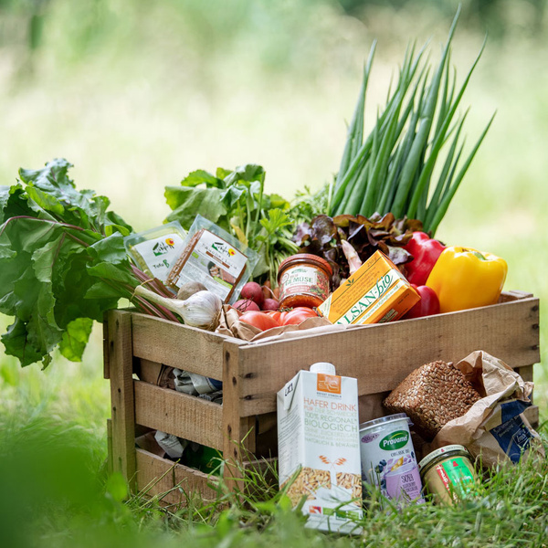 KI generiert: Kiste mit frischem Gemüse, Brot, Kräutern und Bio-Produkten auf einer Wiese. Text: "Hafer Drink 100% regional - biologisch".
