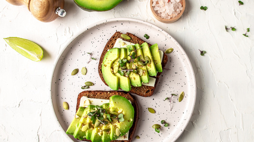 Zwei Brotscheiben mit Avocado und Microgreens auf einem Teller, daneben Limetten, Salz und Pfeffer.