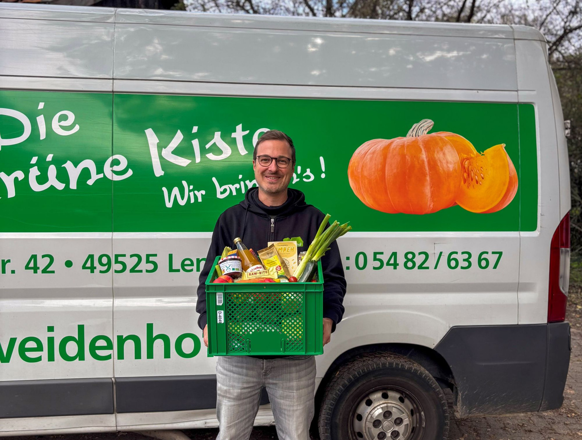 KI generiert: Ein Mann hält eine Kiste mit Lebensmitteln vor einem grünen Lieferwagen mit Kürbis-Aufdruck. Text: „Die grüne Kiste“.