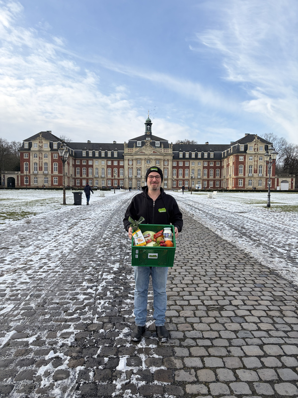KI generiert: Ein Mann hält eine Kiste mit Lebensmitteln vor einem großen Gebäude. Schneebedeckter Boden und klarer Himmel.