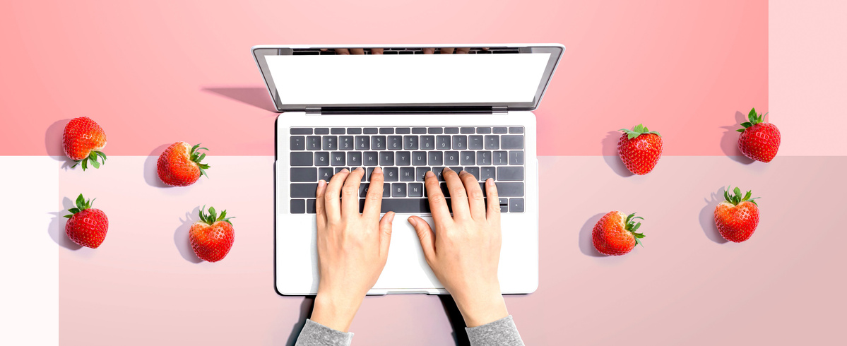 Hände tippen auf einem Laptop, umgeben von Erdbeeren auf rosa Hintergrund.