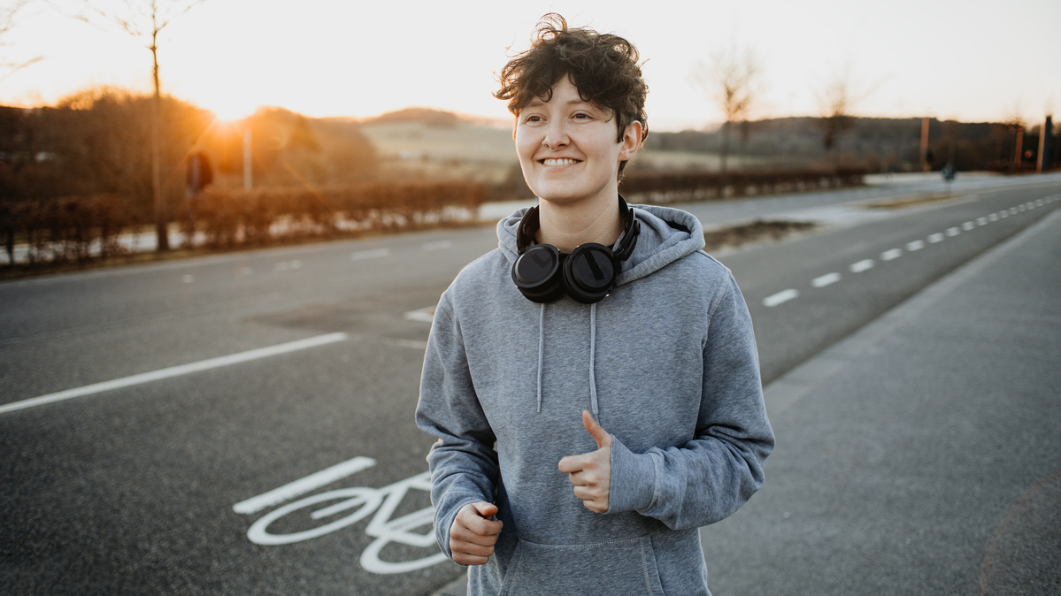 Eine Person joggt auf einem Radweg mit Kopfhörern am Hals, im Hintergrund Sonnenuntergang.