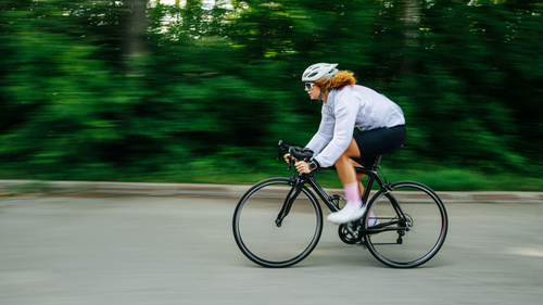 Eine Person fährt schnell mit einem Rennrad auf einer Straße, hinter grüner, unscharfer Landschaft.