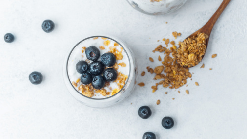 Joghurt mit Heidelbeeren und Müsli in einem Glas, daneben verstreutes Müsli.