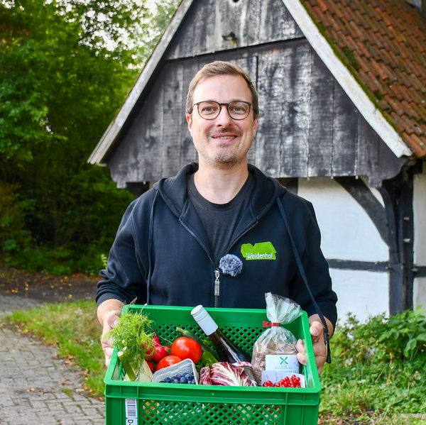 KI generiert: Ein Mann hält eine Kiste mit frischen Lebensmitteln. Text: "Weidenhof".