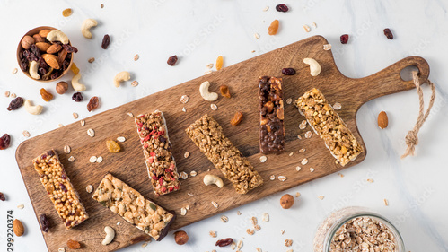 Mehrere Müsli- und Nussriegel auf einem Holzbrett, umgeben von Nüssen und Haferflocken.