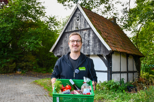 KI generiert: Ein Mann hält eine Kiste mit frischem Gemüse vor einem Fachwerkhäuschen.