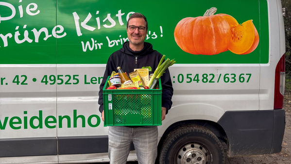 Ein Mann hält eine grüne Kiste mit Lebensmitteln vor einem Lieferwagen mit buntem Schriftzug und Kürbis.  Text: "Die grüne Kiste Wir bringen's!"