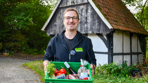KI generiert: Eine Person hält eine grüne Kiste mit frischem Gemüse vor einem traditionellen Haus. Text: "Weidenhof".