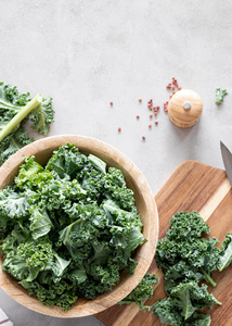 Eine Schüssel mit frischem Grünkohl, einem Schneidebrett und Gewürzen auf einer Küchenarbeitsfläche.