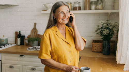 Eine Frau in gelbem Hemd telefoniert und hält eine Tasse in einer gemütlichen Küche.