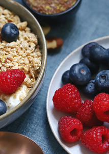 Müsli in einer Schüssel mit Joghurt und Beeren, daneben ein Teller mit Himbeeren und Blaubeeren.