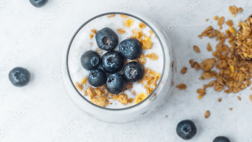Joghurt mit Heidelbeeren und Müsli in einem Glas, daneben verstreutes Müsli.